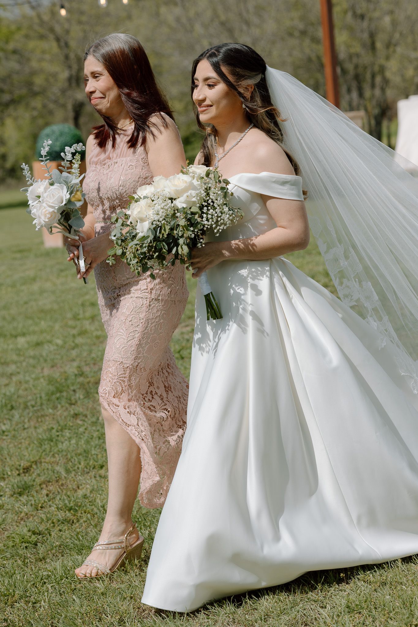 Bride in off-shoulder gown and bridesmaid in pink lace dress hold white bouquets outdoors.