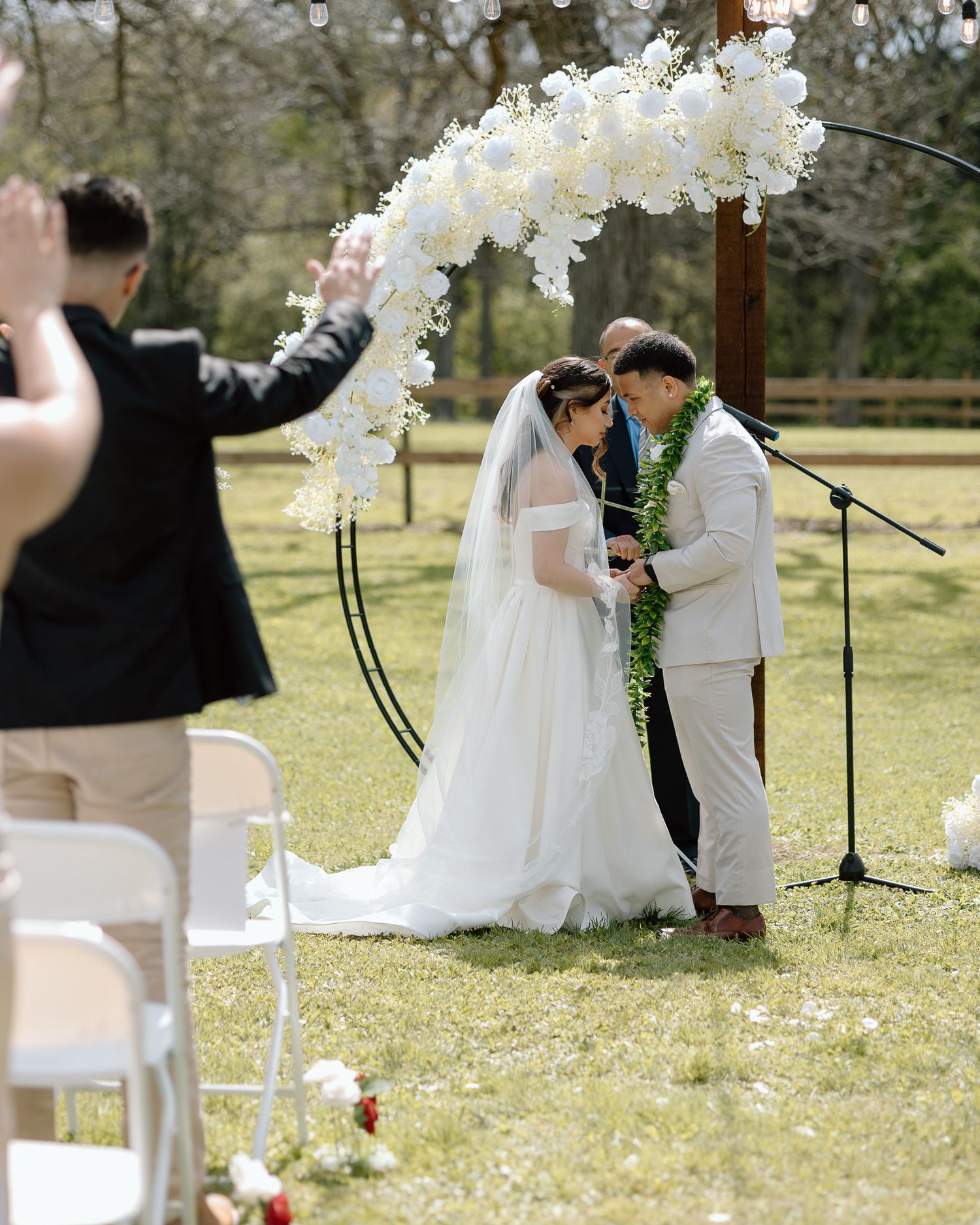 An outdoor wedding ceremony under a white floral arch with guests watching the exchange of vows.
