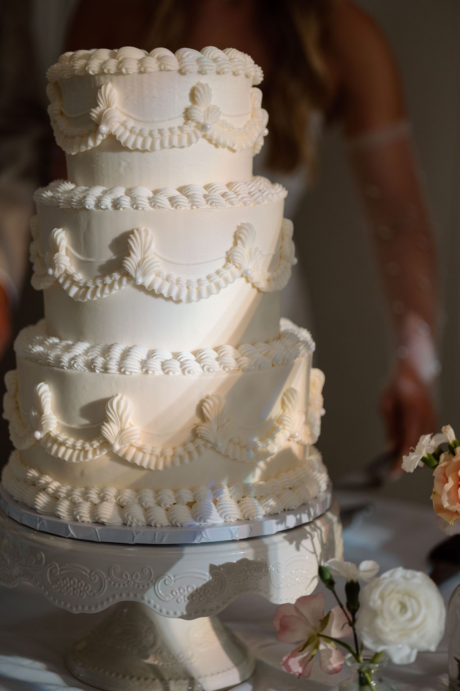 Elegant three-tiered white wedding cake with intricate piped details and seashell decorations on a silver cake stand.