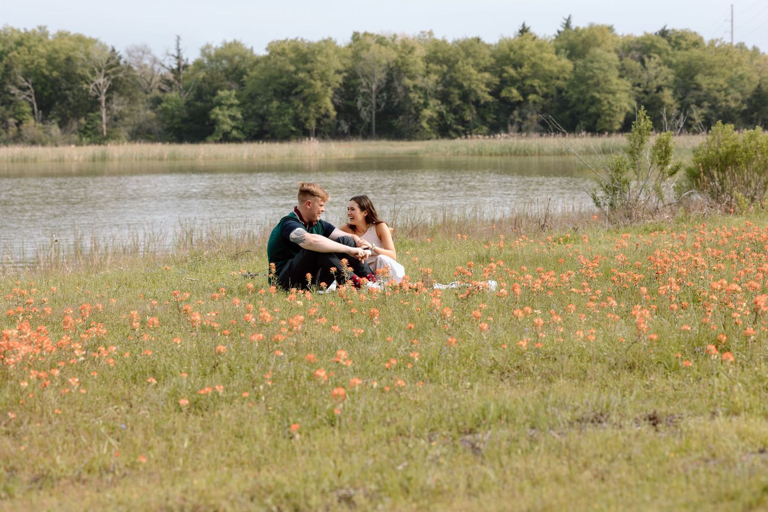 A couple sits together on a grassy hillside surrounded by orange wildflowers near a lake.