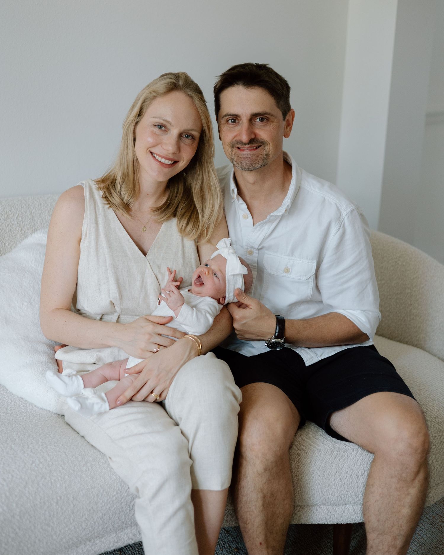 Family portrait on white couch with newborn baby in bright, minimalist setting.