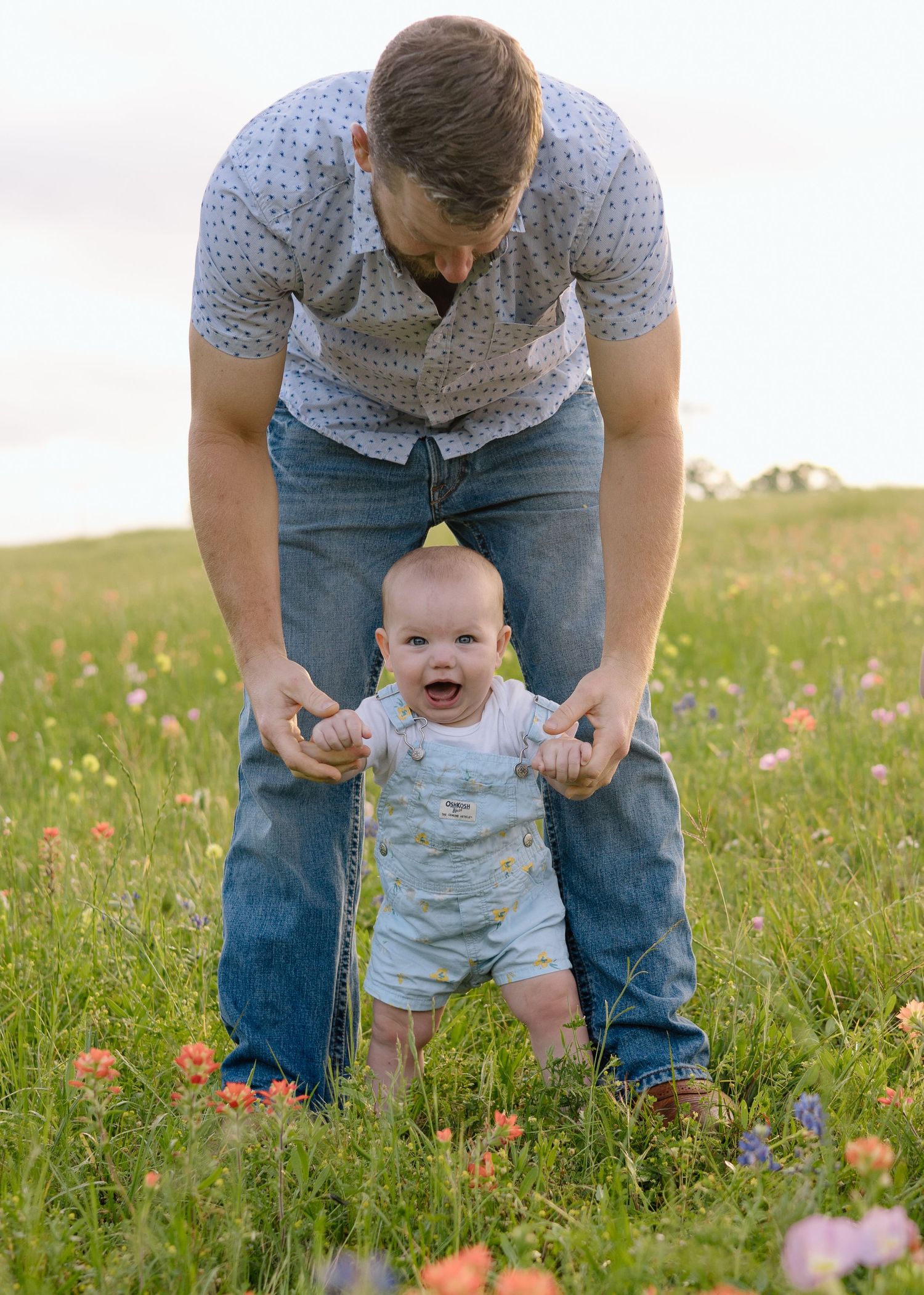 Baby wearing blue overalls takes first steps in a wildflower meadow with support during sunset.