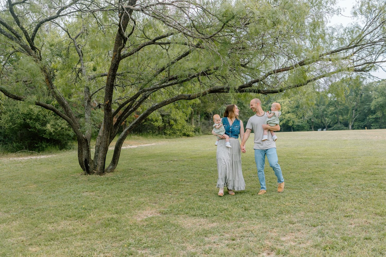 Two people stand chatting together in a grassy park area with a large tree in the background.