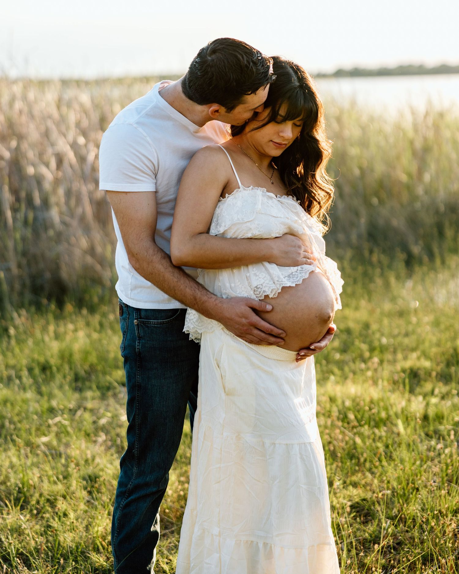 Couple embracing outdoors in golden light, pregnant woman in white dress with partner in white tee and jeans in field.