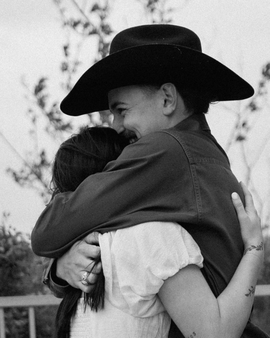 Black and white photo of a couple embracing, man wearing a cowboy hat hugging a woman in a white dress outdoors.