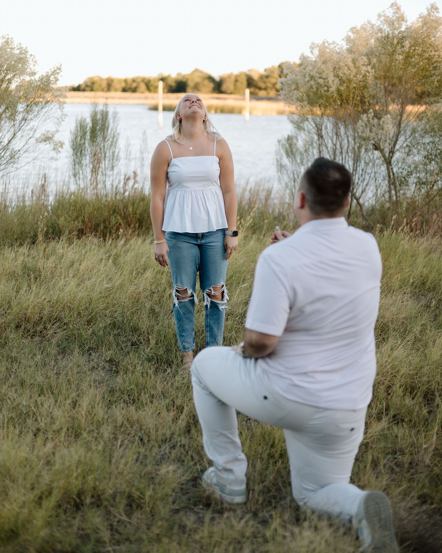 Man kneeling on one knee proposing to a woman near a lake at sunset, both dressed in white and jeans.