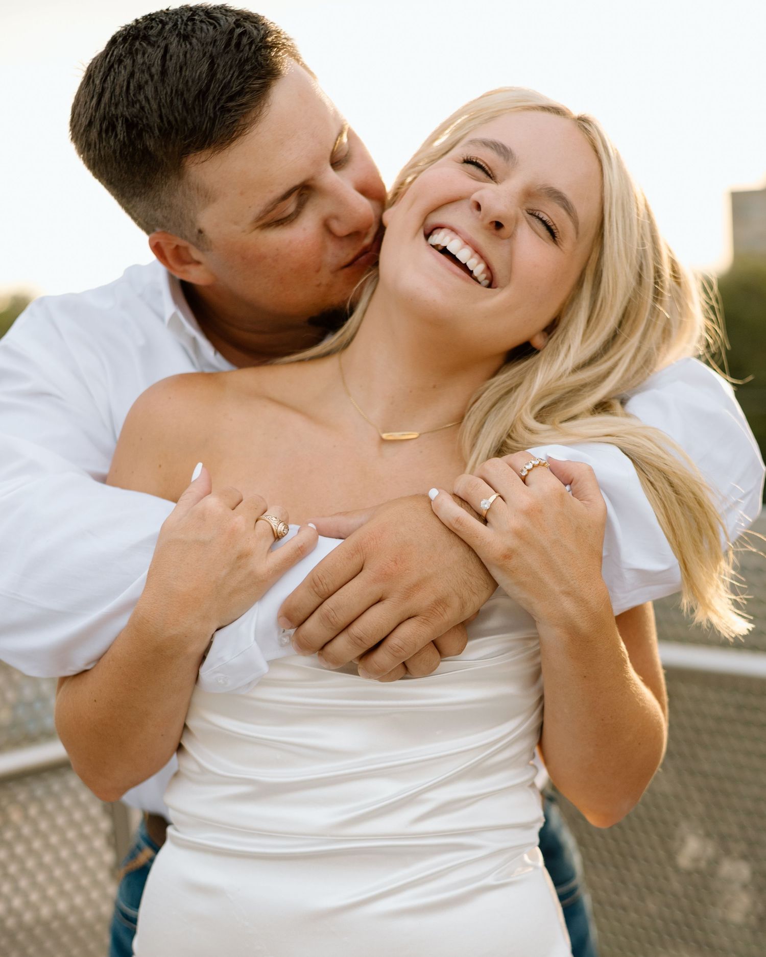 A joyful couple in white outfits share a tender moment, the man kissing the laughing blonde woman's cheek outdoors.