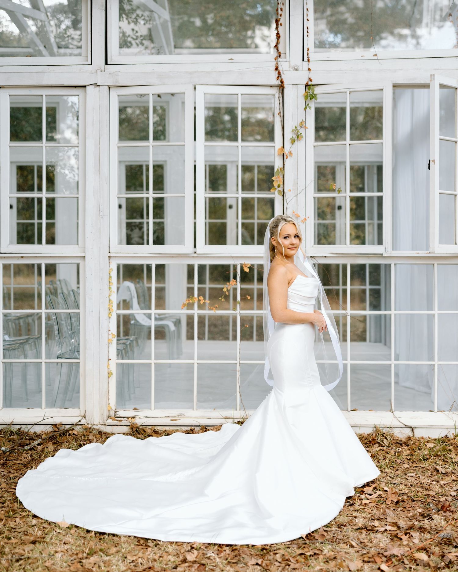 Bride in a strapless mermaid wedding gown with long train and veil poses before white greenhouse windows.