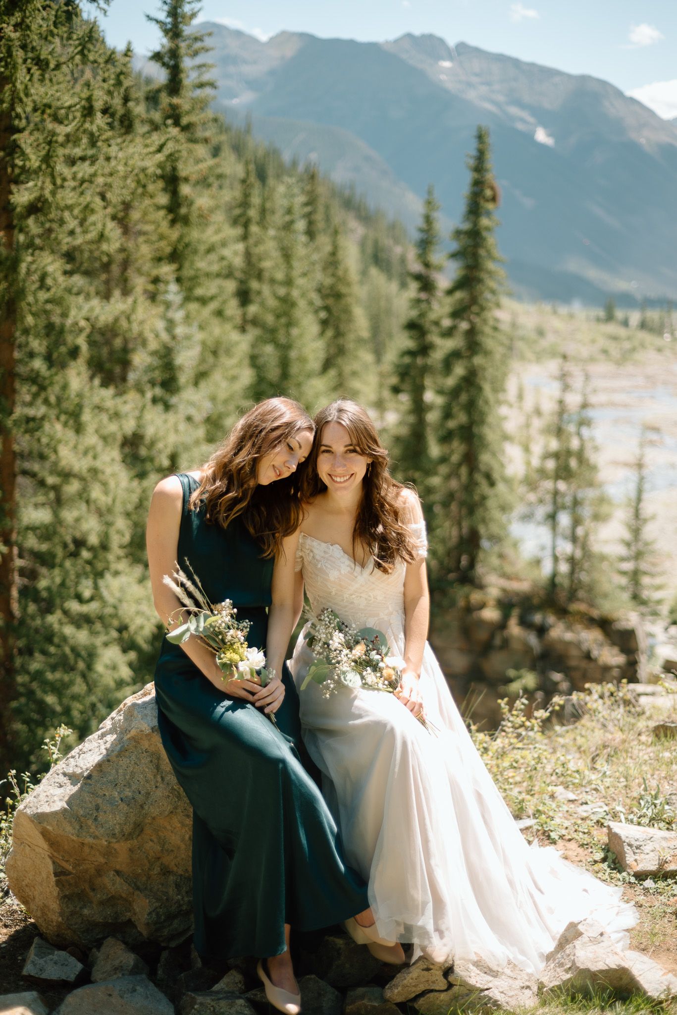 Two figures in elegant dresses share a candid moment on mountain rocks with scenic forest backdrop.