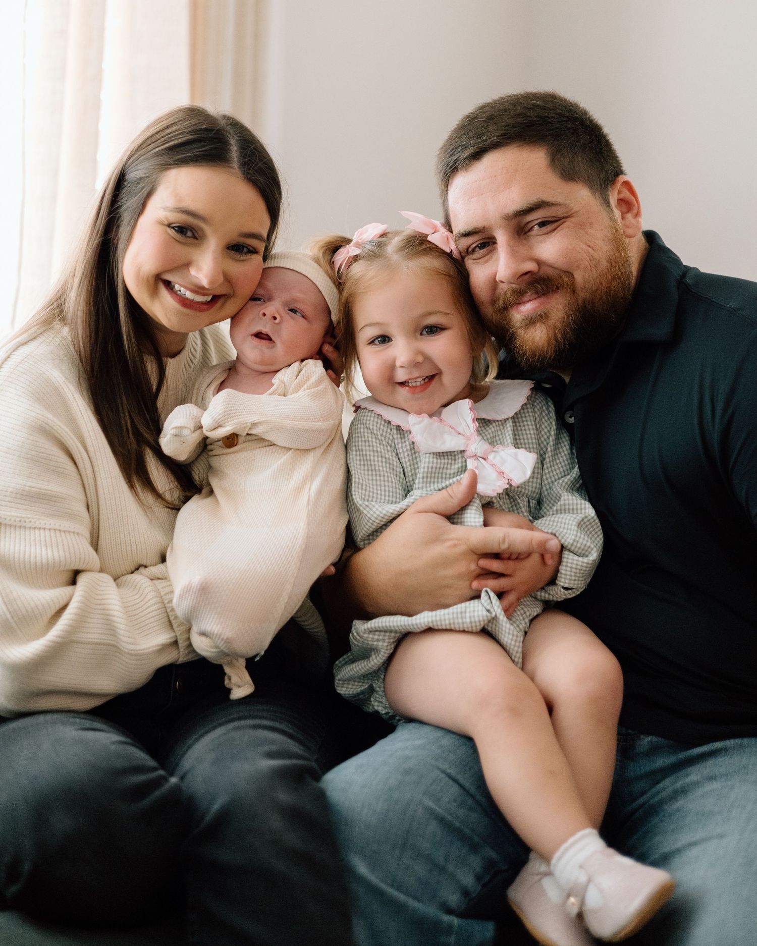 Happy family portrait with parents holding newborn baby and toddler girl with pink bow, sitting together smiling.