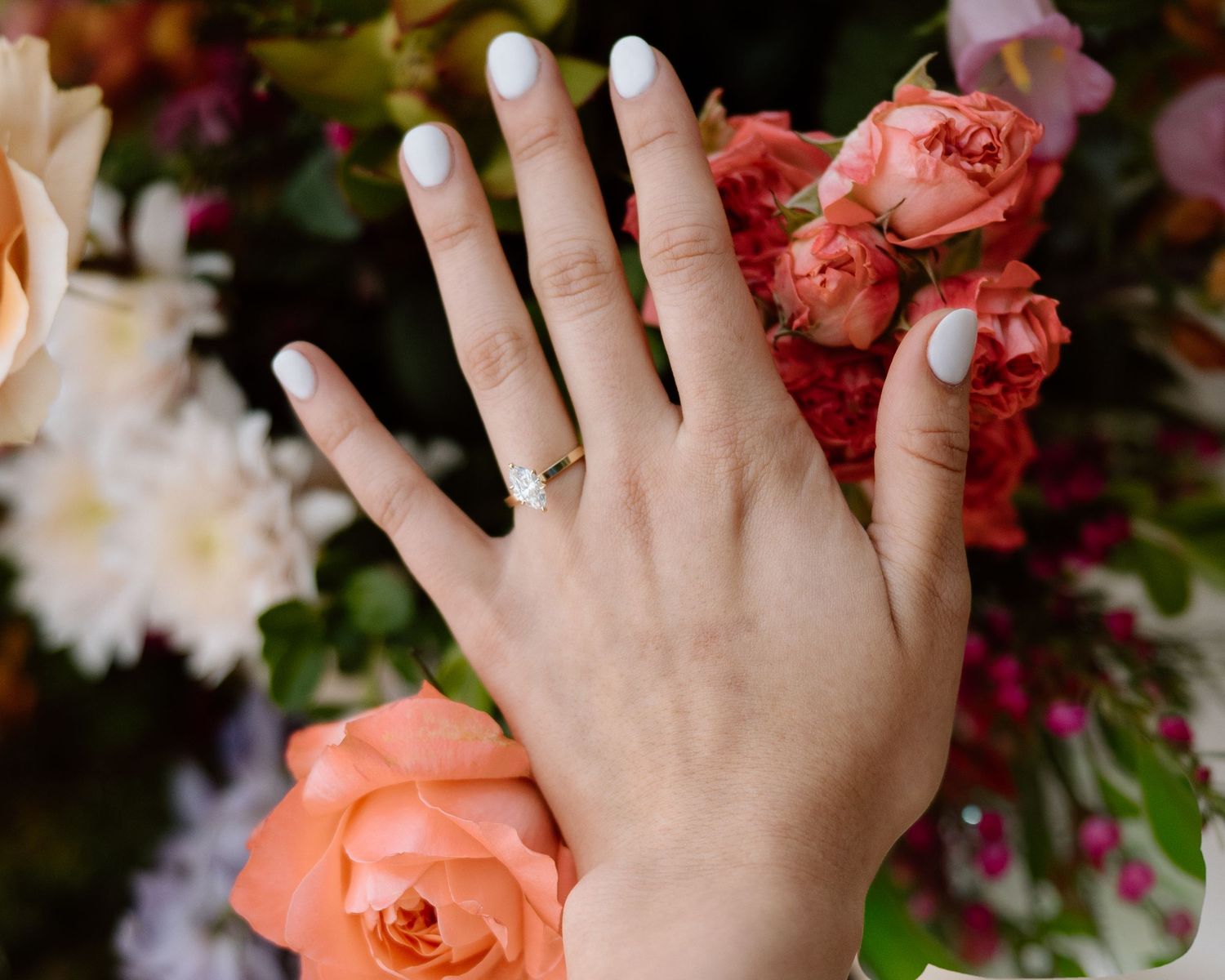 Close-up of a hand with white nails wearing a diamond engagement ring, surrounded by peach roses and flowers.