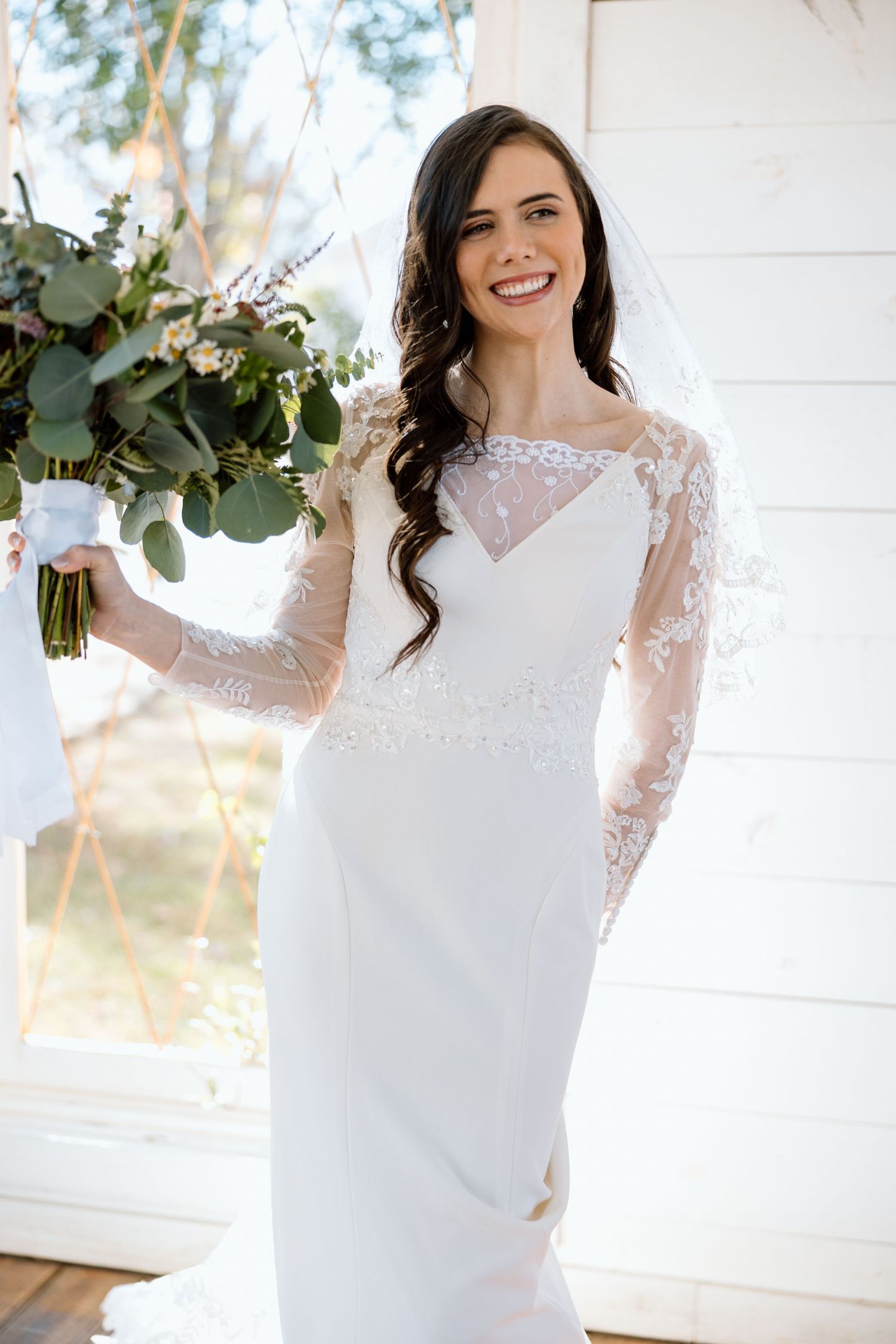 Smiling bride in lace long-sleeve wedding gown holding greenery bouquet outdoors.