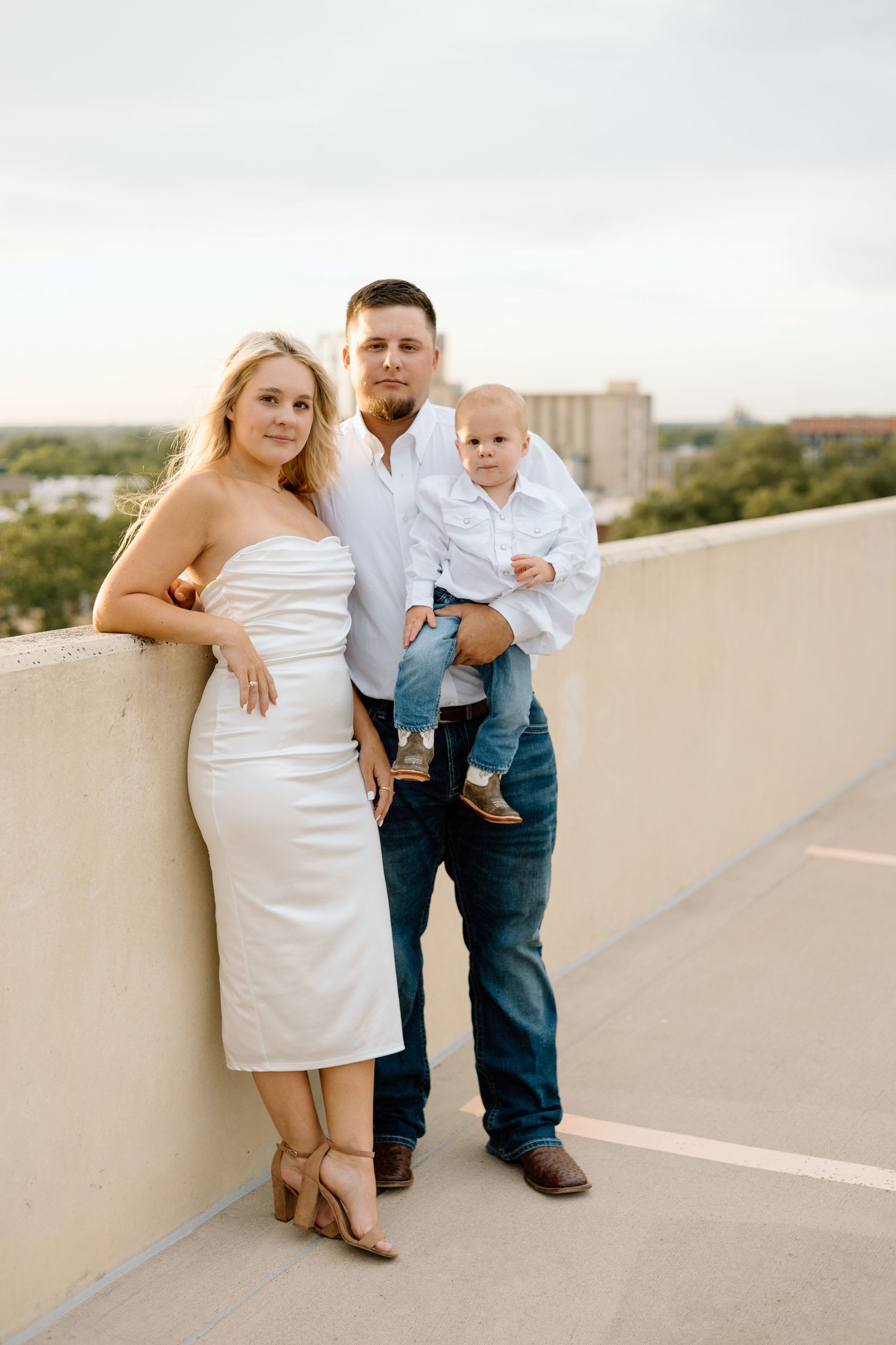 A young family poses on a rooftop at sunset, mother in white dress, father holding toddler, both parents in jeans.