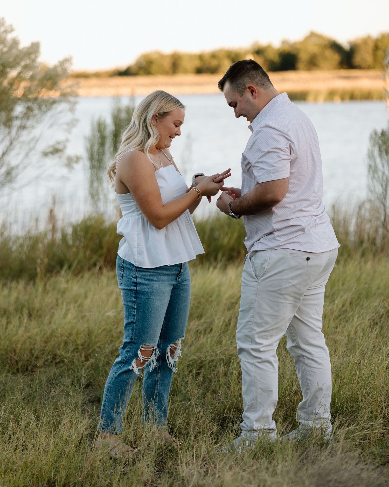 A man places an engagement ring on a woman's finger outdoors near a lake at golden hour.