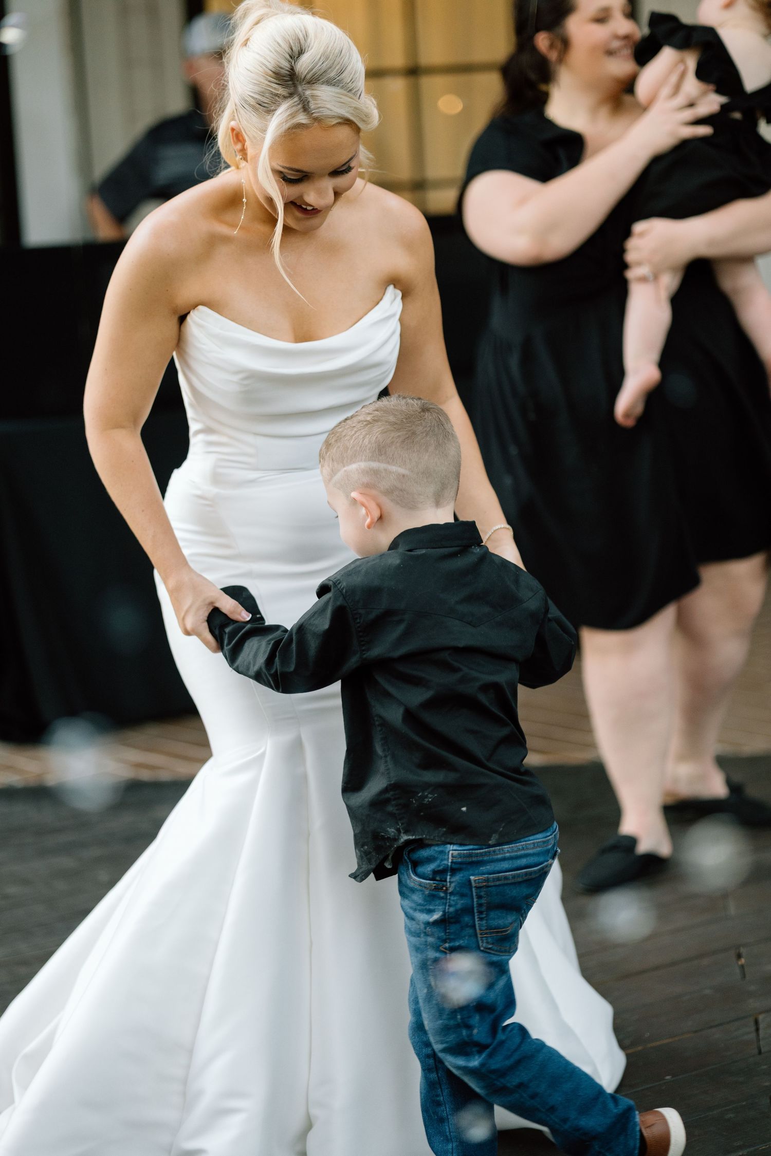 Bride in strapless mermaid gown smiles while dancing with young boy in black shirt at wedding reception.