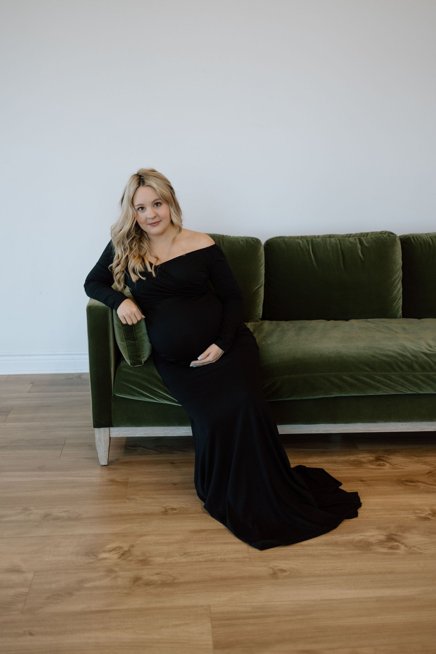 A person in a long black formal gown reclines elegantly on a dark green velvet couch against a white wall.