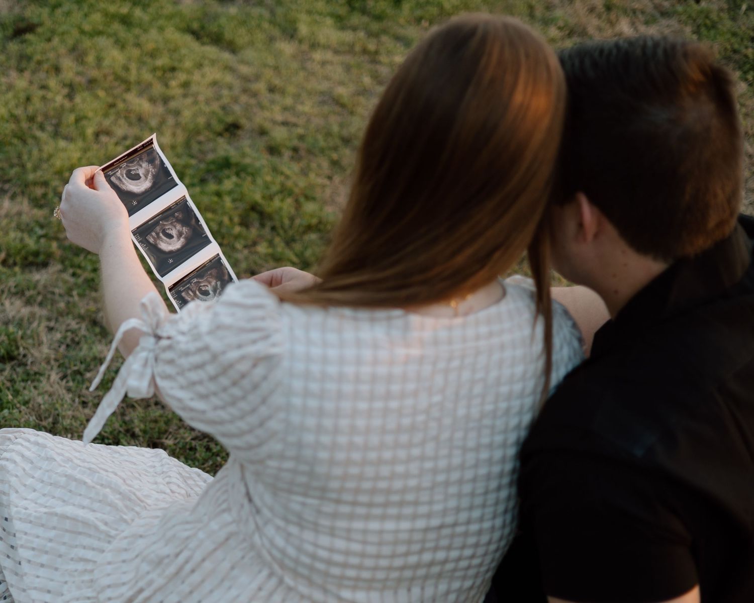 Couple looks at ultrasound photos together while sitting on grassy shore.