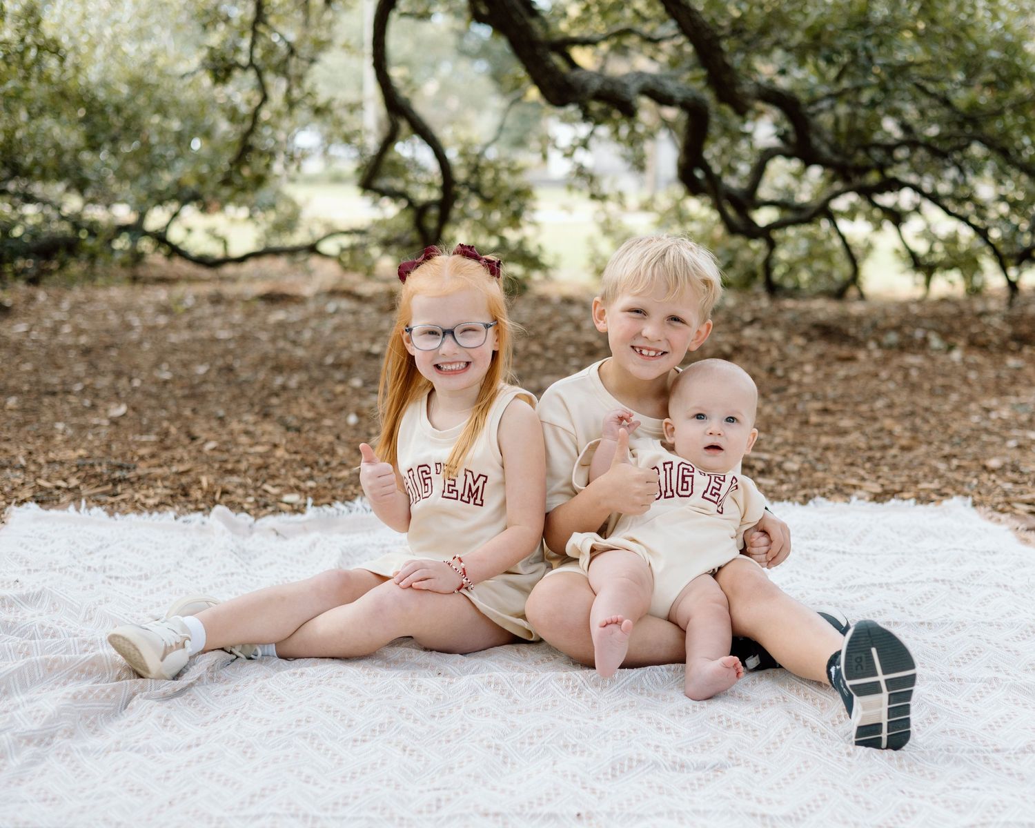 Three young siblings smile together on a white blanket outdoors, wearing matching cream TAMU outfits.