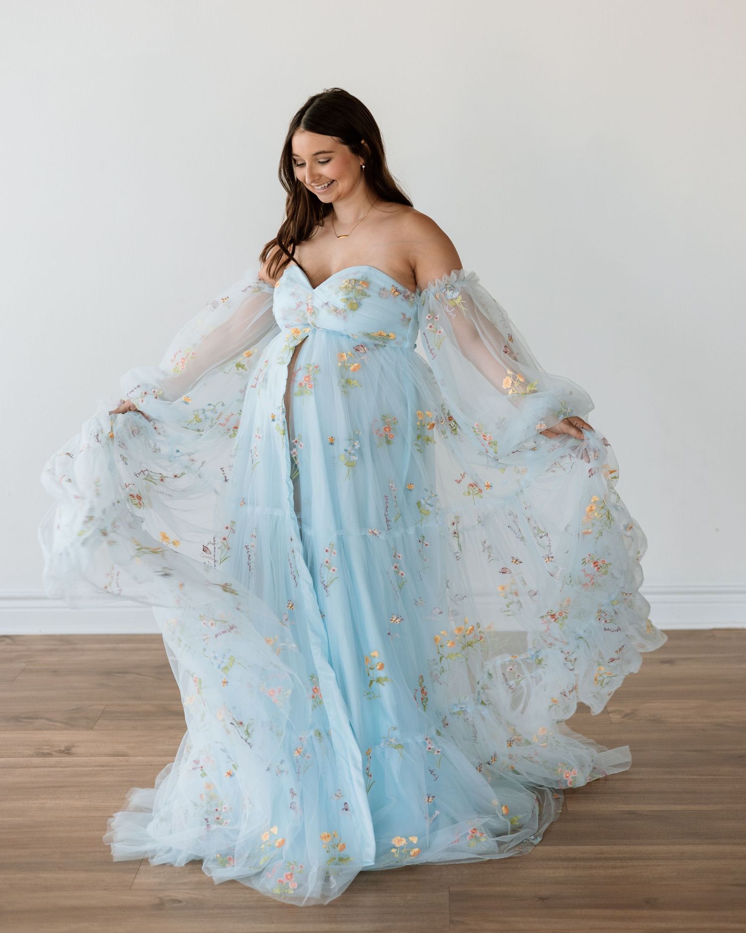 Pregnant woman in a flowing light blue floral tulle off-shoulder gown, posing gracefully in a bright studio.