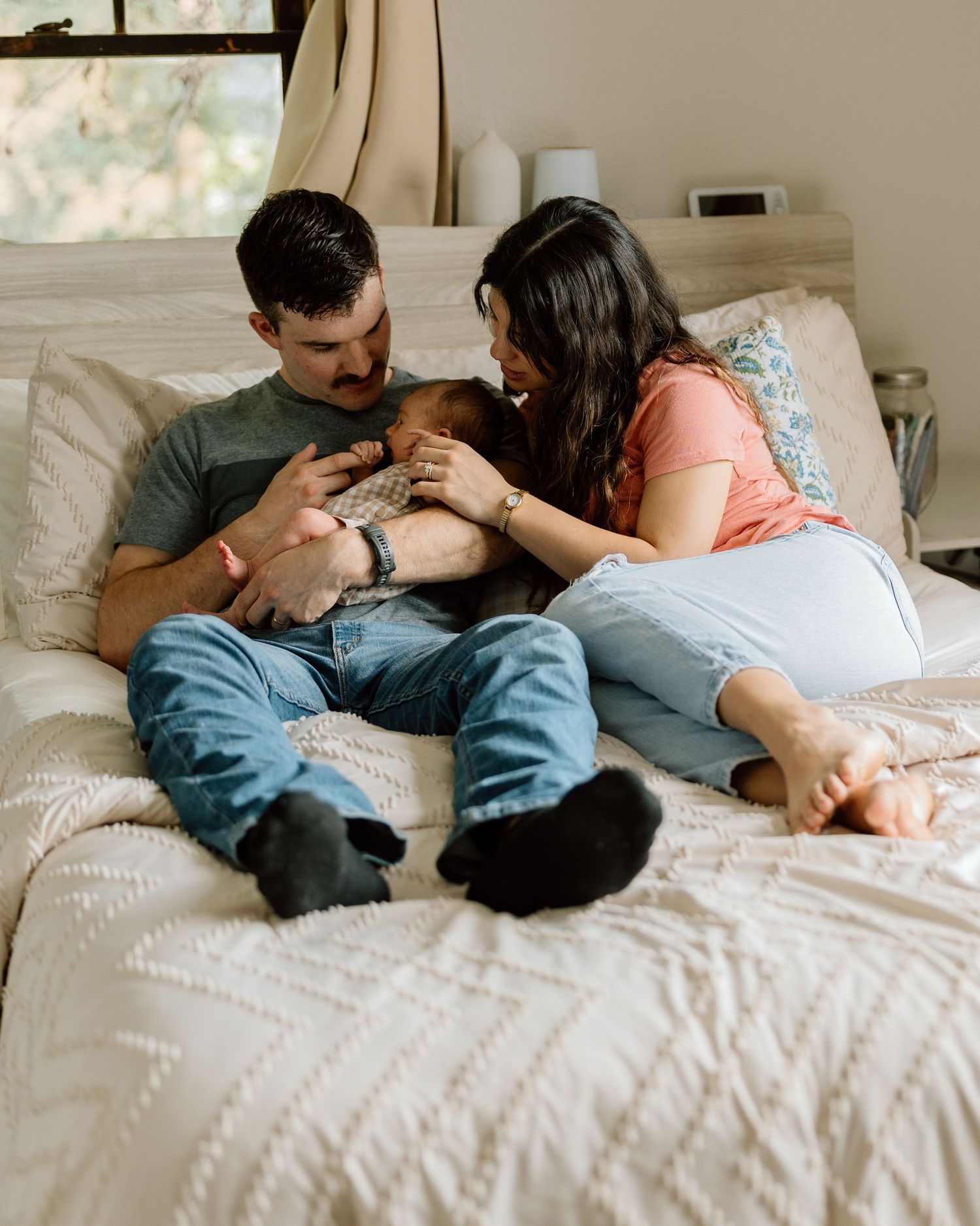 A couple sits on a bed, lovingly cradling their newborn baby in a warm, cozy bedroom setting.
