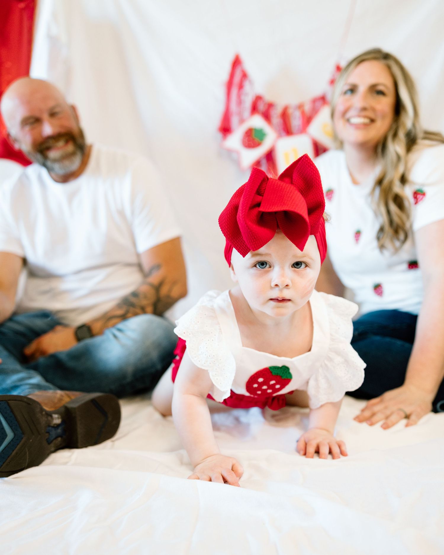 Baby girl in strawberry outfit and red bow headband crawls toward camera as parents smile behind her.