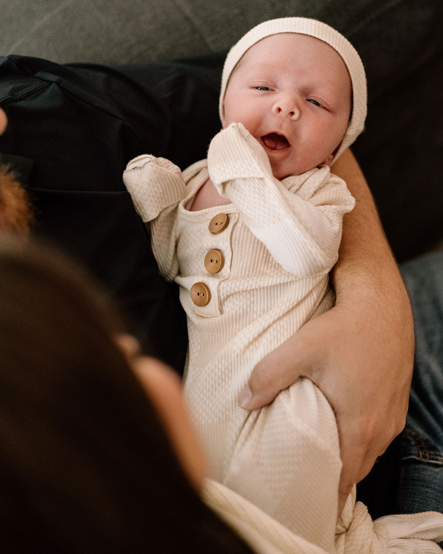 Adorable newborn baby in cream outfit and hat yawning while being held by parent on dark sofa.