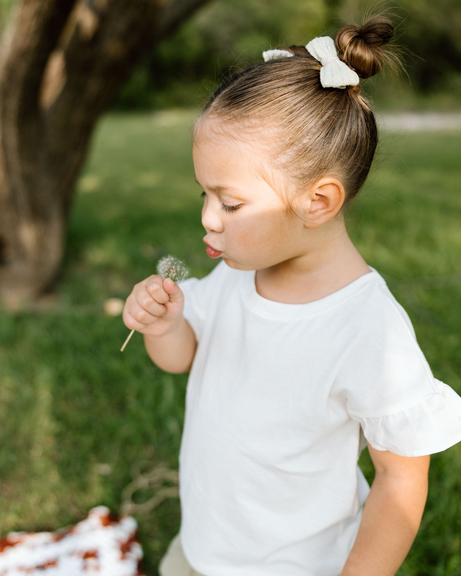 A cute toddler girl with a bun hairstyle blows a dandelion outdoors in a lush green park.