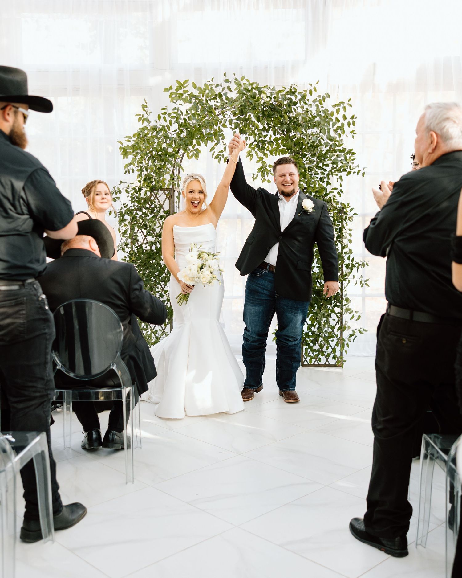 Newlyweds raising hands in celebration while walking down the aisle beneath a lush greenery arch.