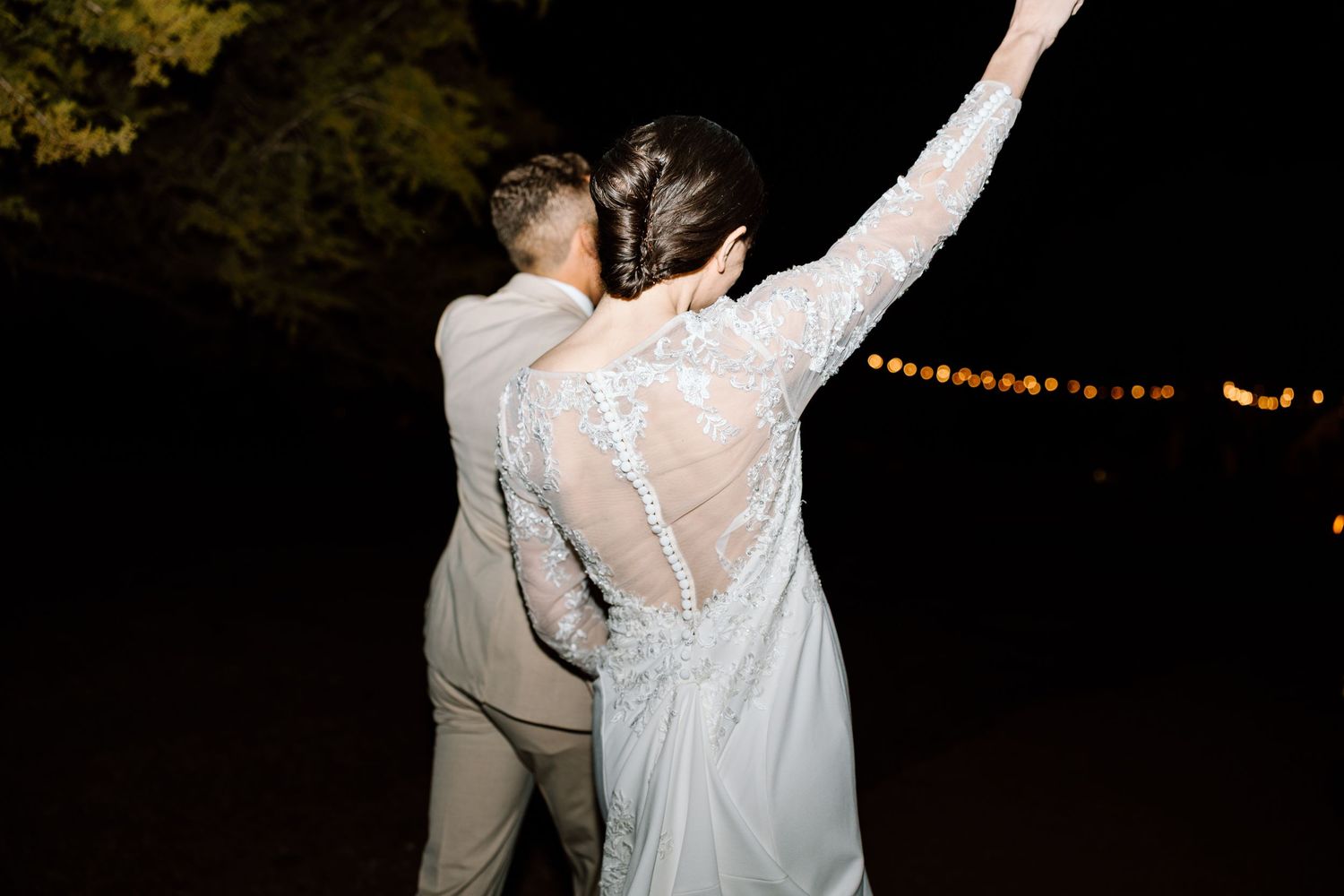 Bride in lace back wedding gown raises arm joyfully as groom embraces her during nighttime celebration.