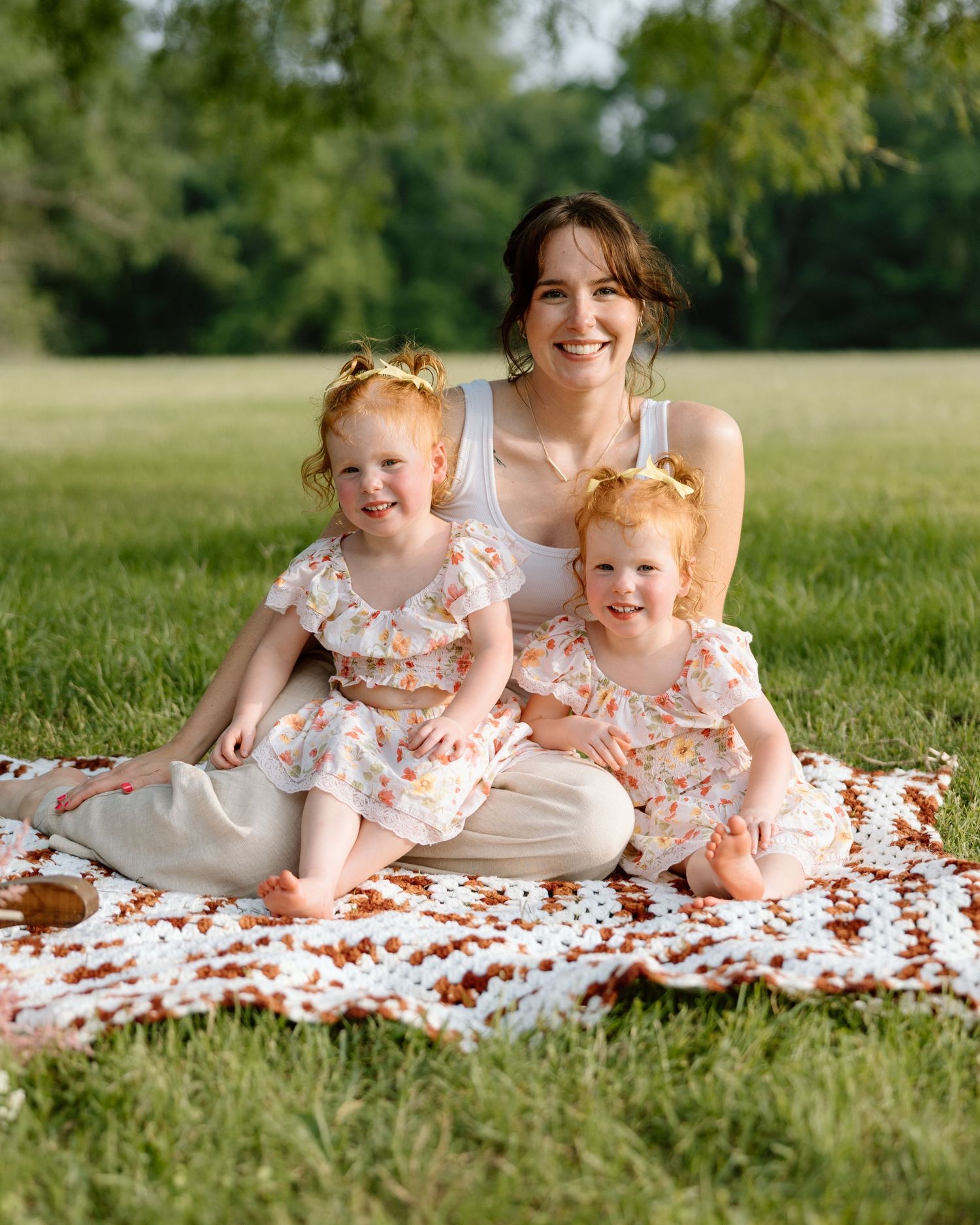 A mother sits on a floral blanket in a green park with her two young redheaded daughters in matching floral dresses.