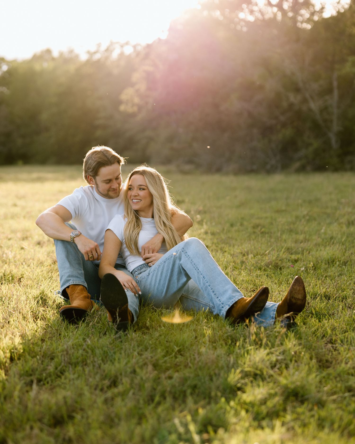 Couple sitting together on green grass field at golden hour sunset, wearing jeans and boots, smiling warmly.