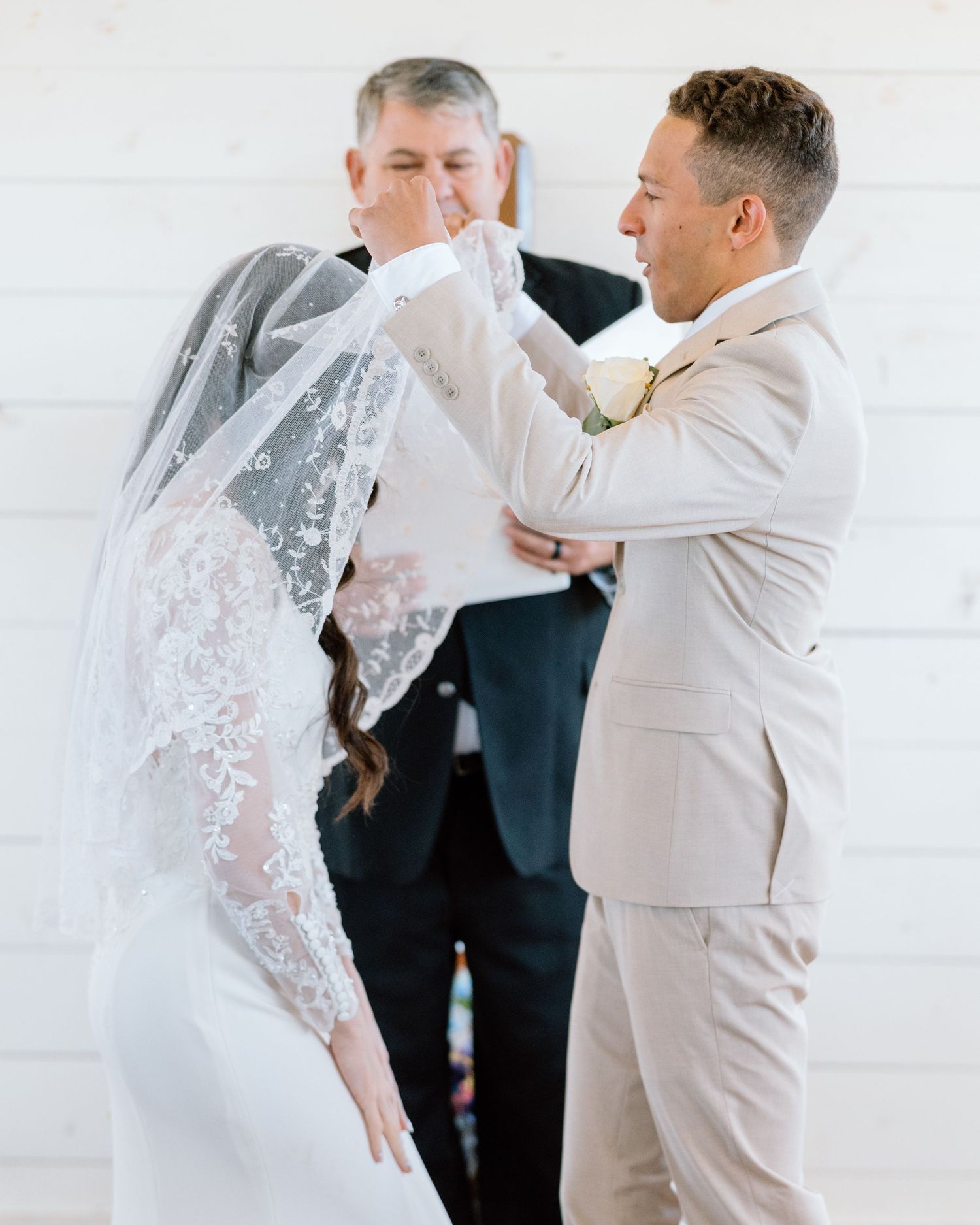 Groom lifting bride's lace veil during wedding ceremony at a white barn venue.