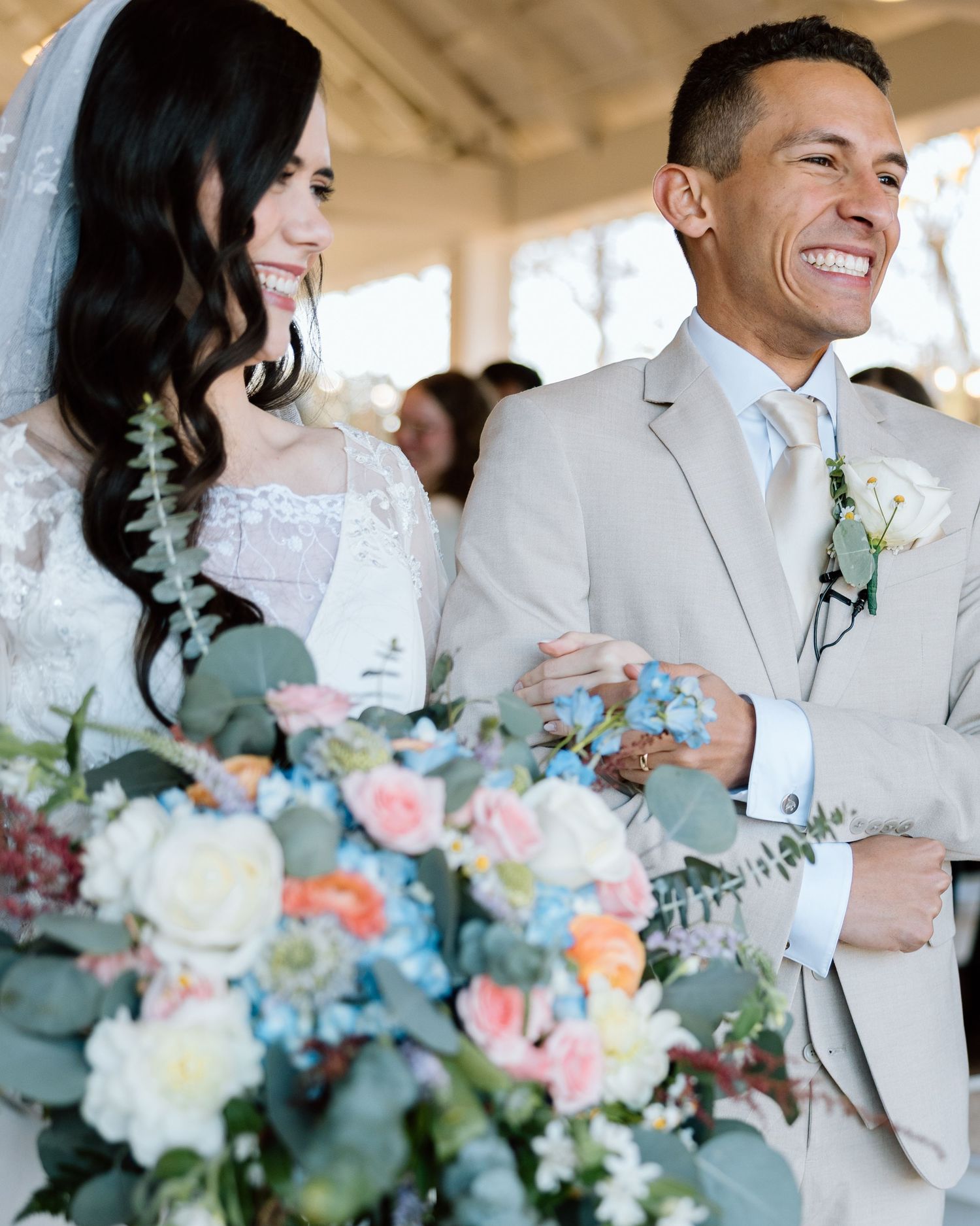 Smiling bride and groom stand together during outdoor wedding ceremony, bride holding colorful bouquet.
