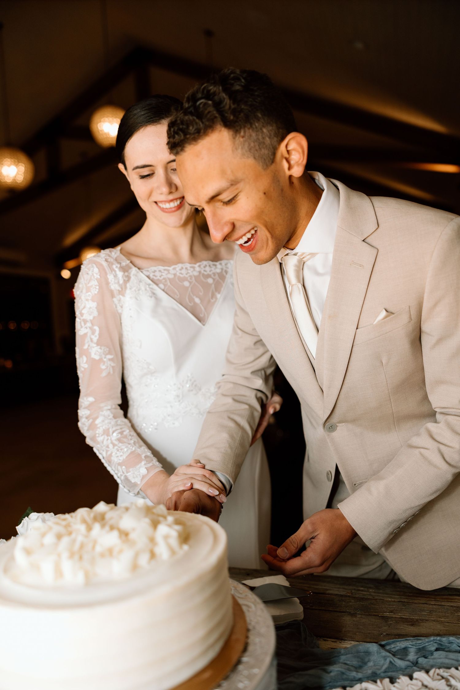 Smiling newlyweds cutting white wedding cake together, groom in beige suit and bride in lace gown.