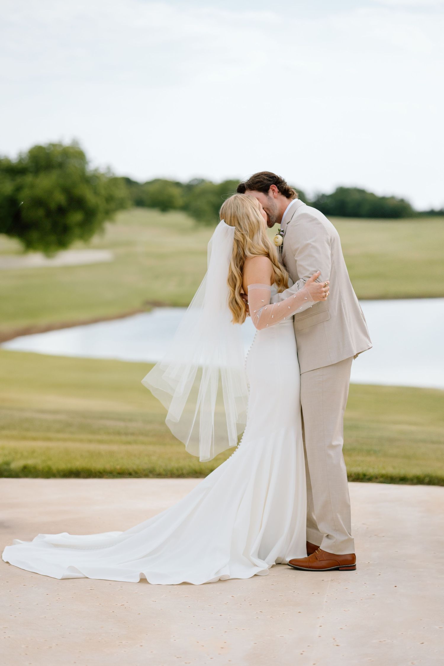 A romantic wedding couple sharing a kiss on a golf course with a lake in the background.
