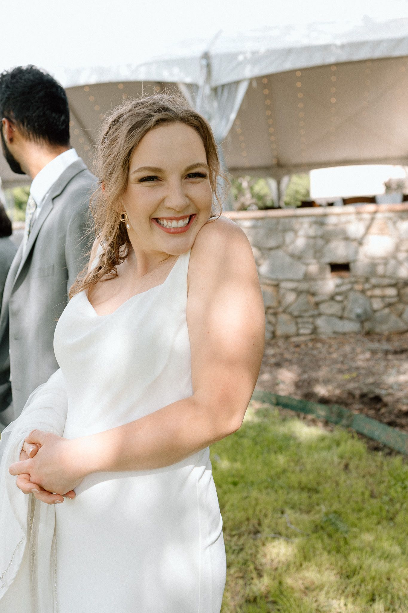 Elegant bride in white dress smiles joyfully under outdoor wedding tent with stone wall backdrop.