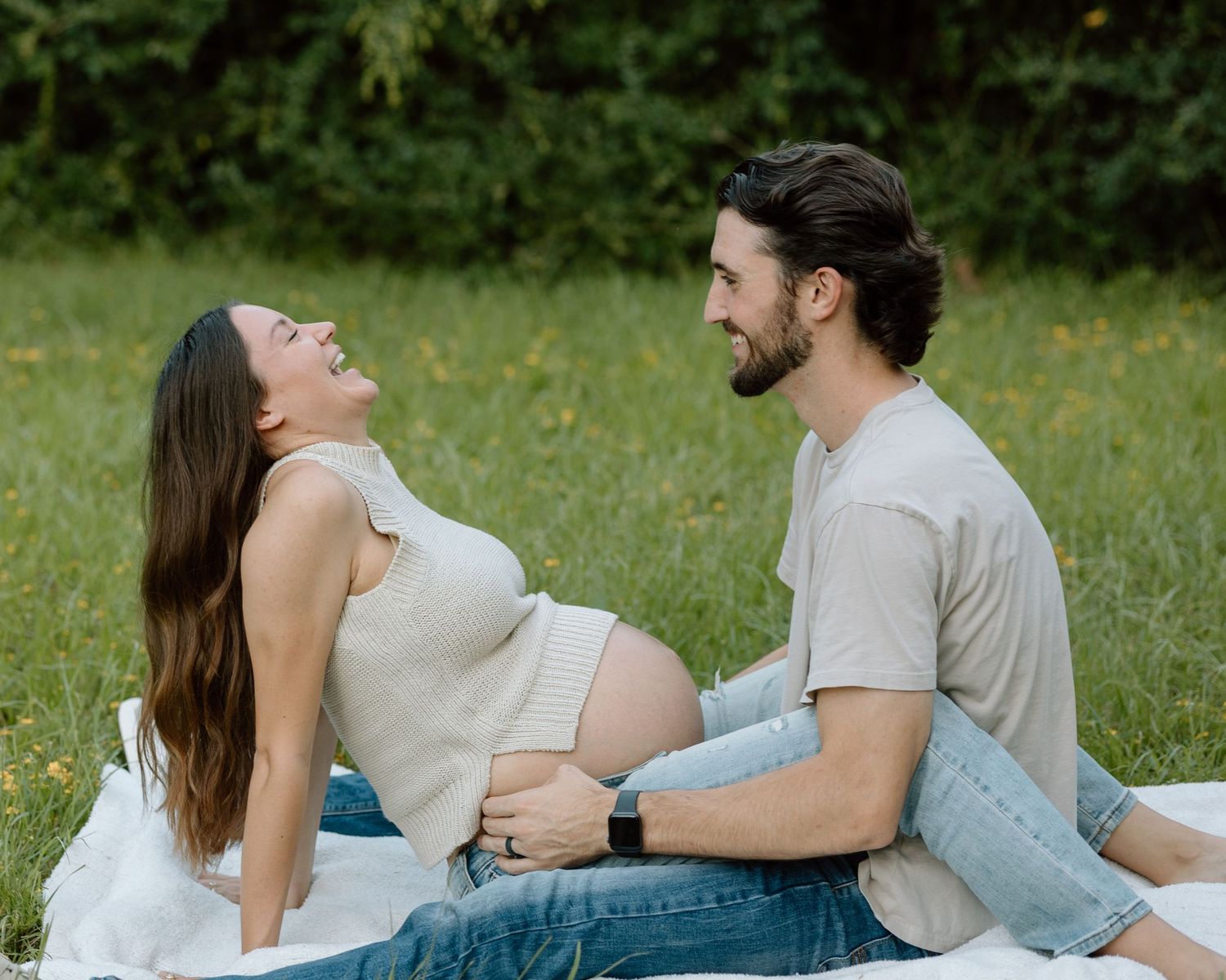 A couple shares laughter during an outdoor maternity photoshoot in a grassy field.