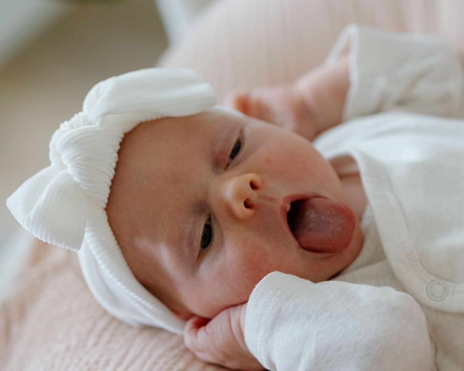 Sleepy baby in white outfit and bow headband yawning while lying down.