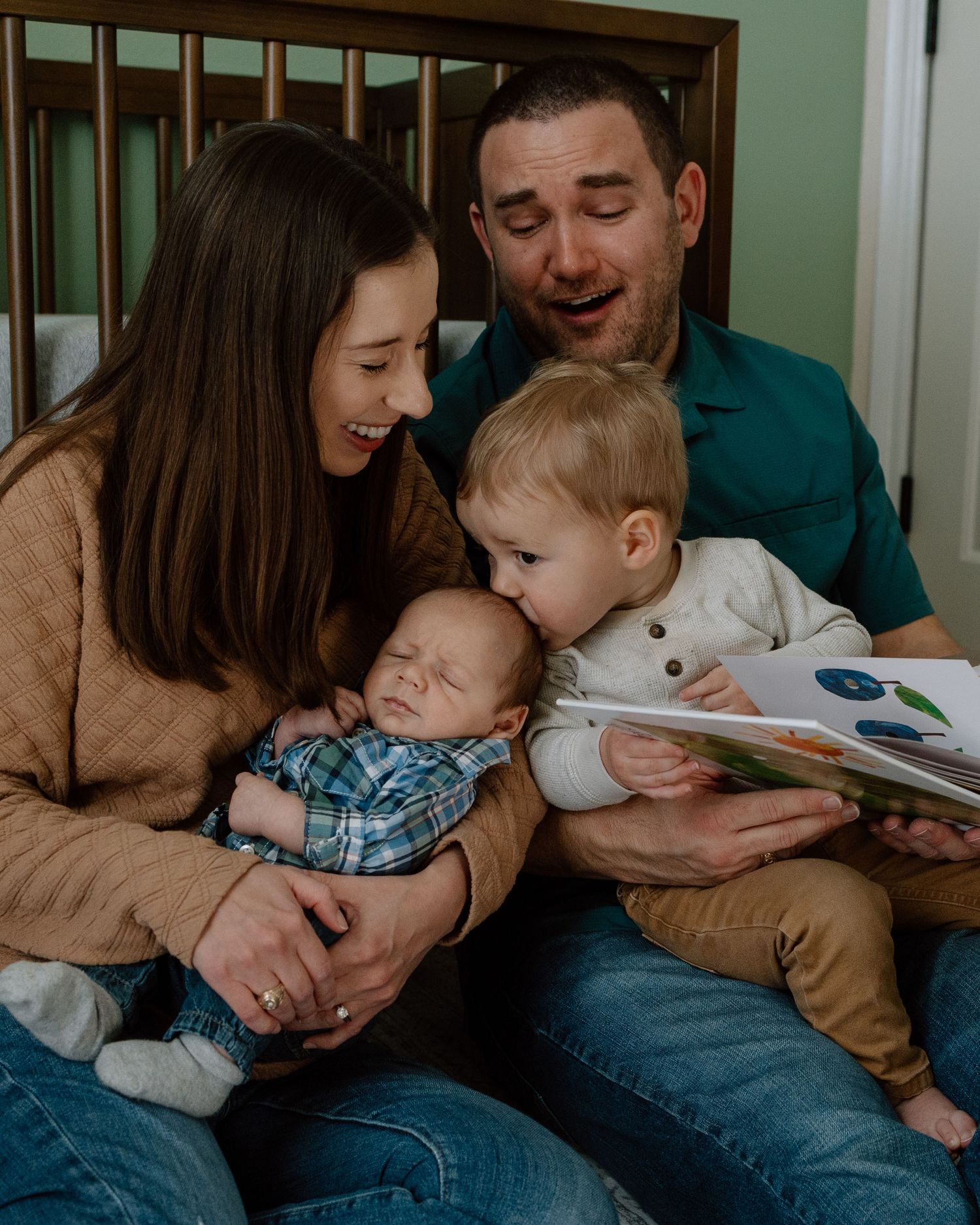 Family sits together on floor reading a children's book while snuggling with a sleeping baby.