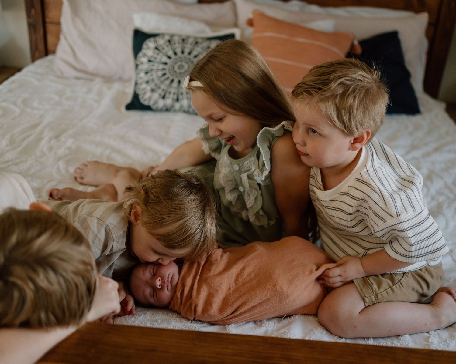 Children in casual clothing play together and share laughter on a cozy bed with decorative pillows.