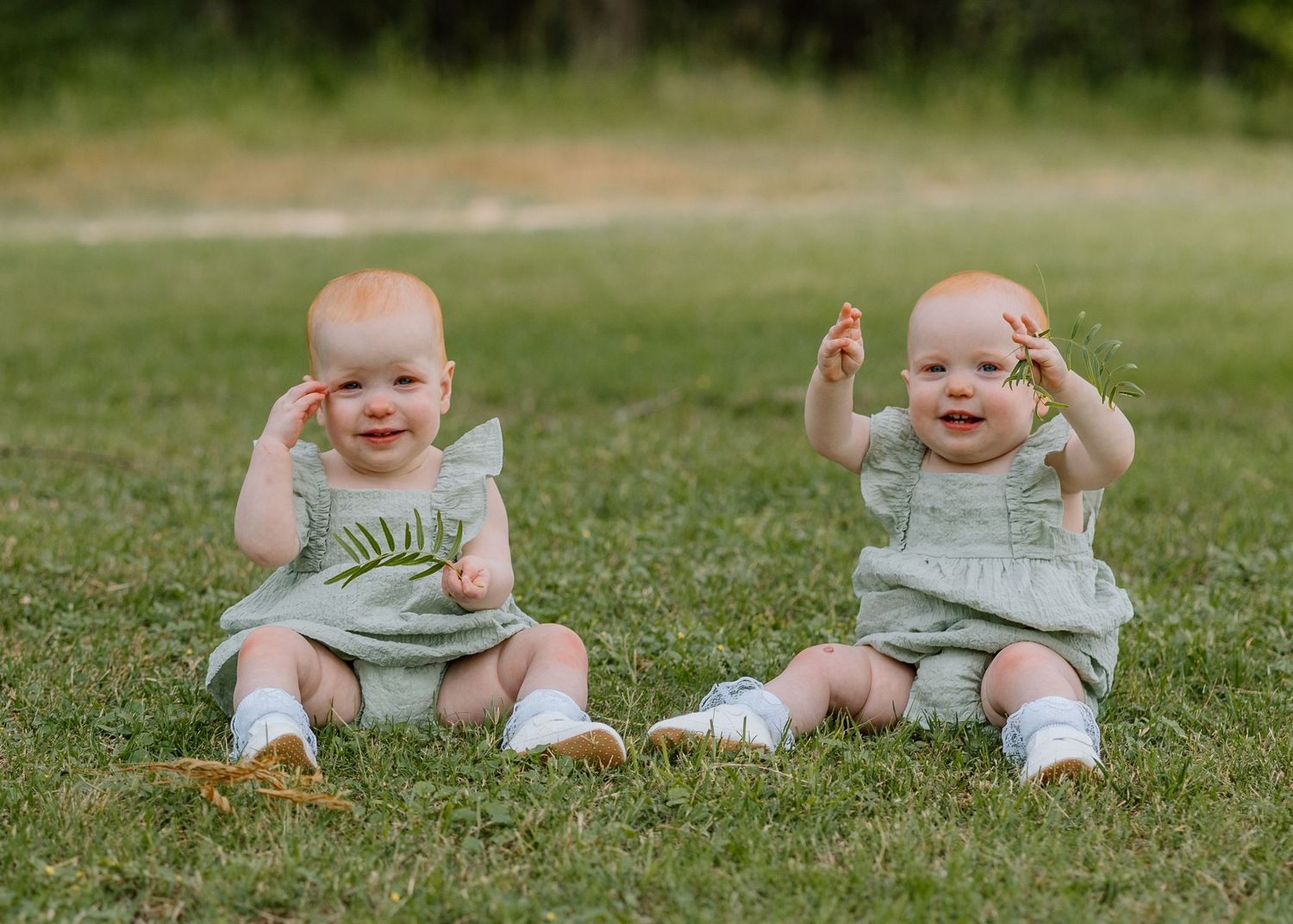 Two small infants in matching light-colored outfits sit together on grass while happily waving their hands.