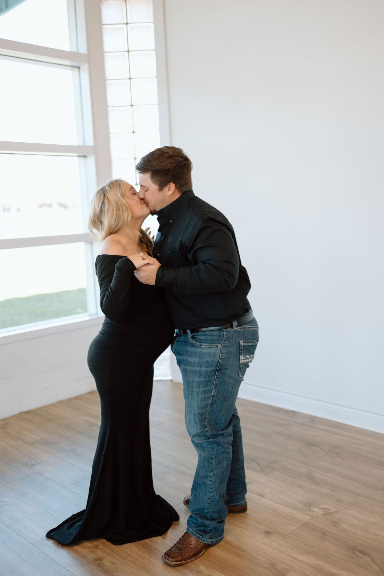 A romantic couple embraces in a bright room with hardwood floors, the woman wearing a long black dress.