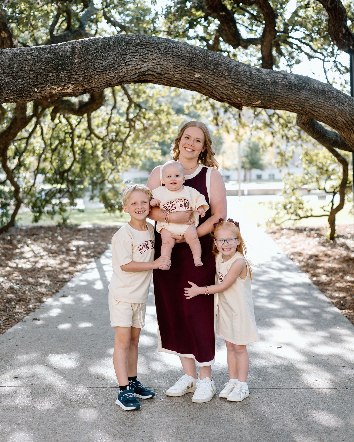 A woman in a maroon dress holds a baby while two young children stand beside her on a shaded park path.