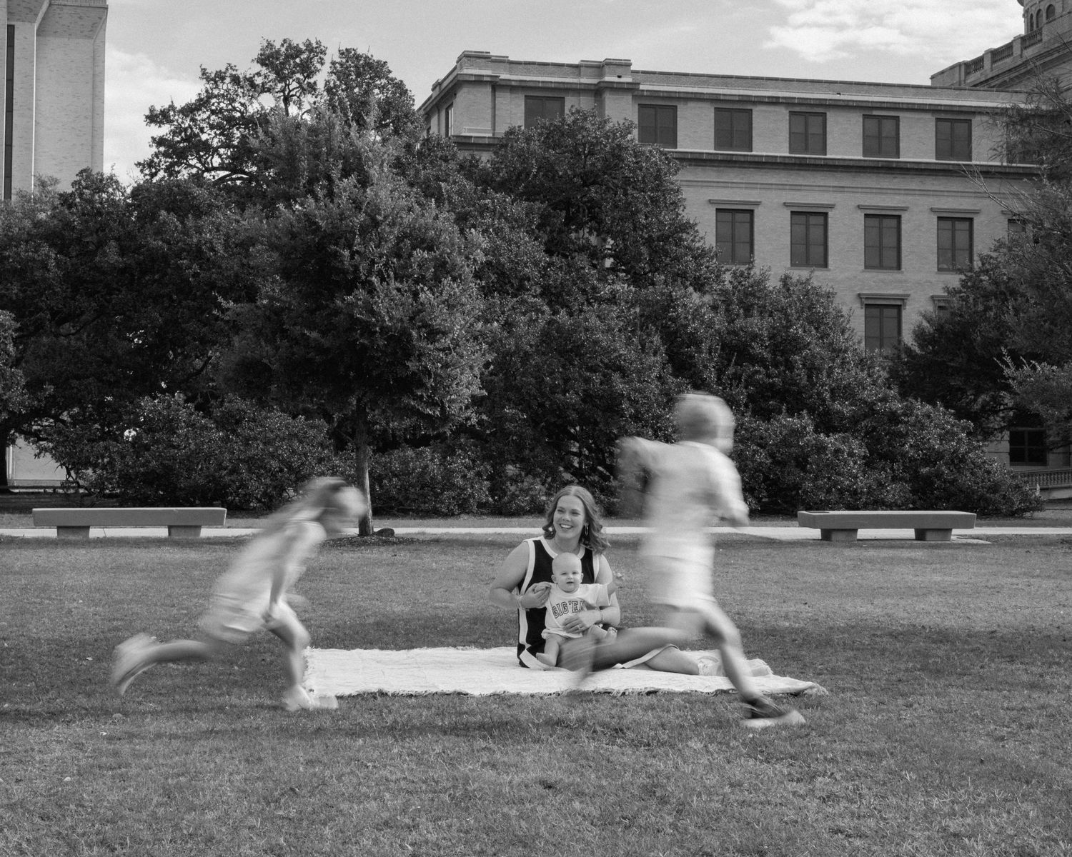 Black and white photo of a woman sitting on grass while two children run past her near a historic building.