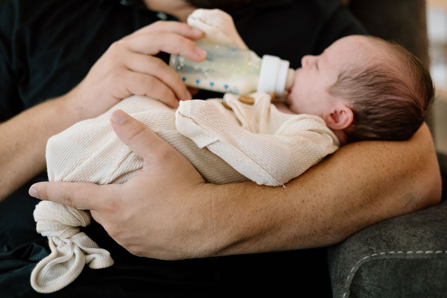Parent cradling a newborn baby while bottle feeding, close-up of infant in white onesie being held securely.