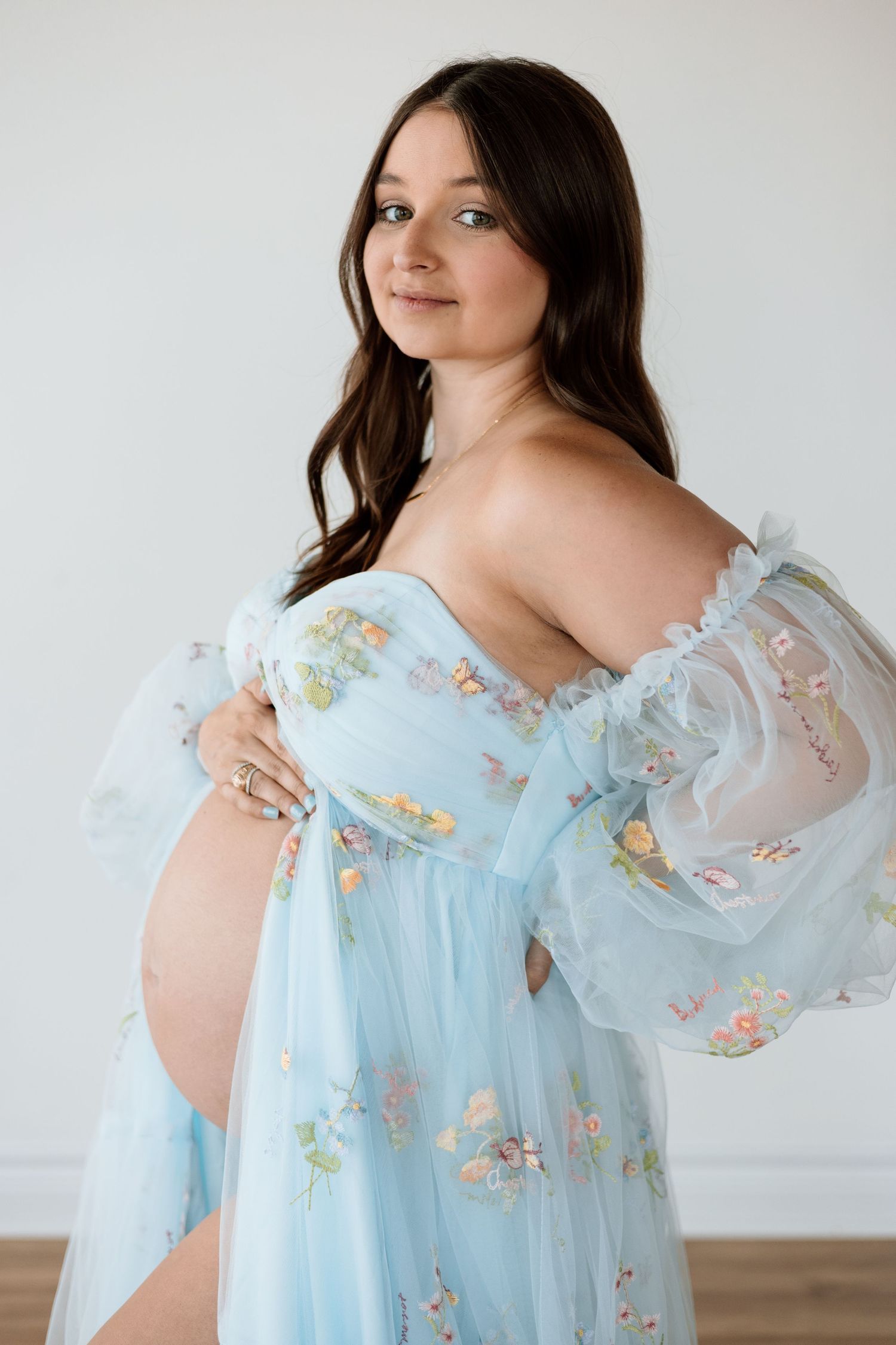 Pregnant woman in floral blue tulle maternity gown cradling baby bump in bright white photography studio.