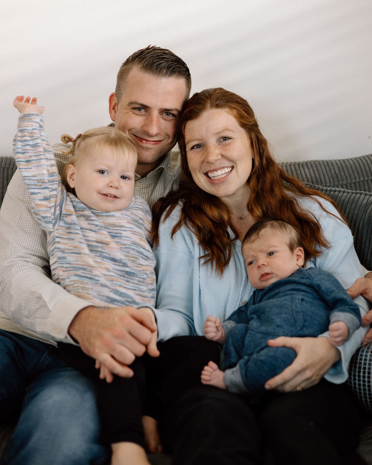 Happy family of four with two young children smiling together on a couch, including a newborn baby in blue.