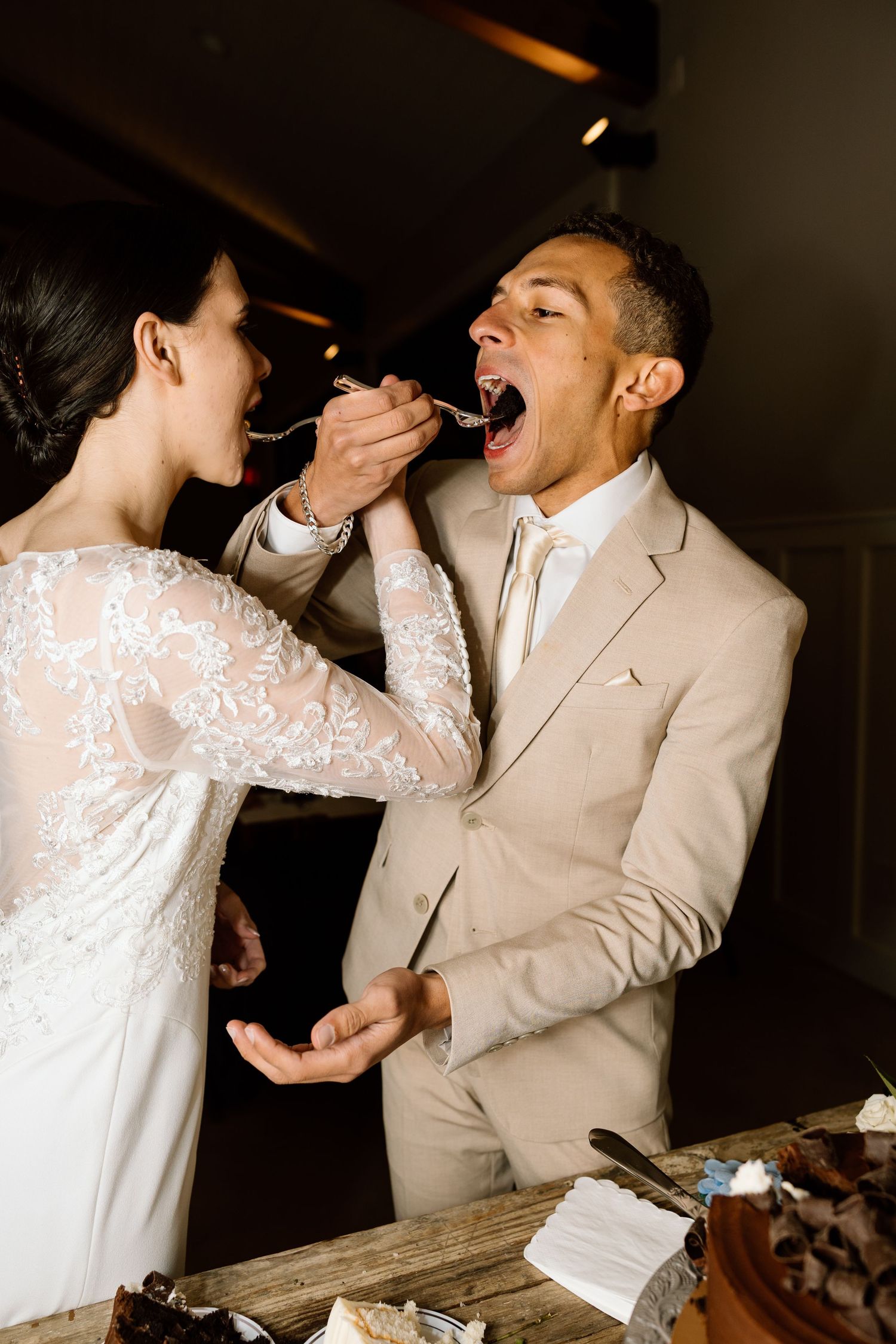Bride in lace gown feeds wedding cake to groom in beige suit during romantic cake cutting ceremony.