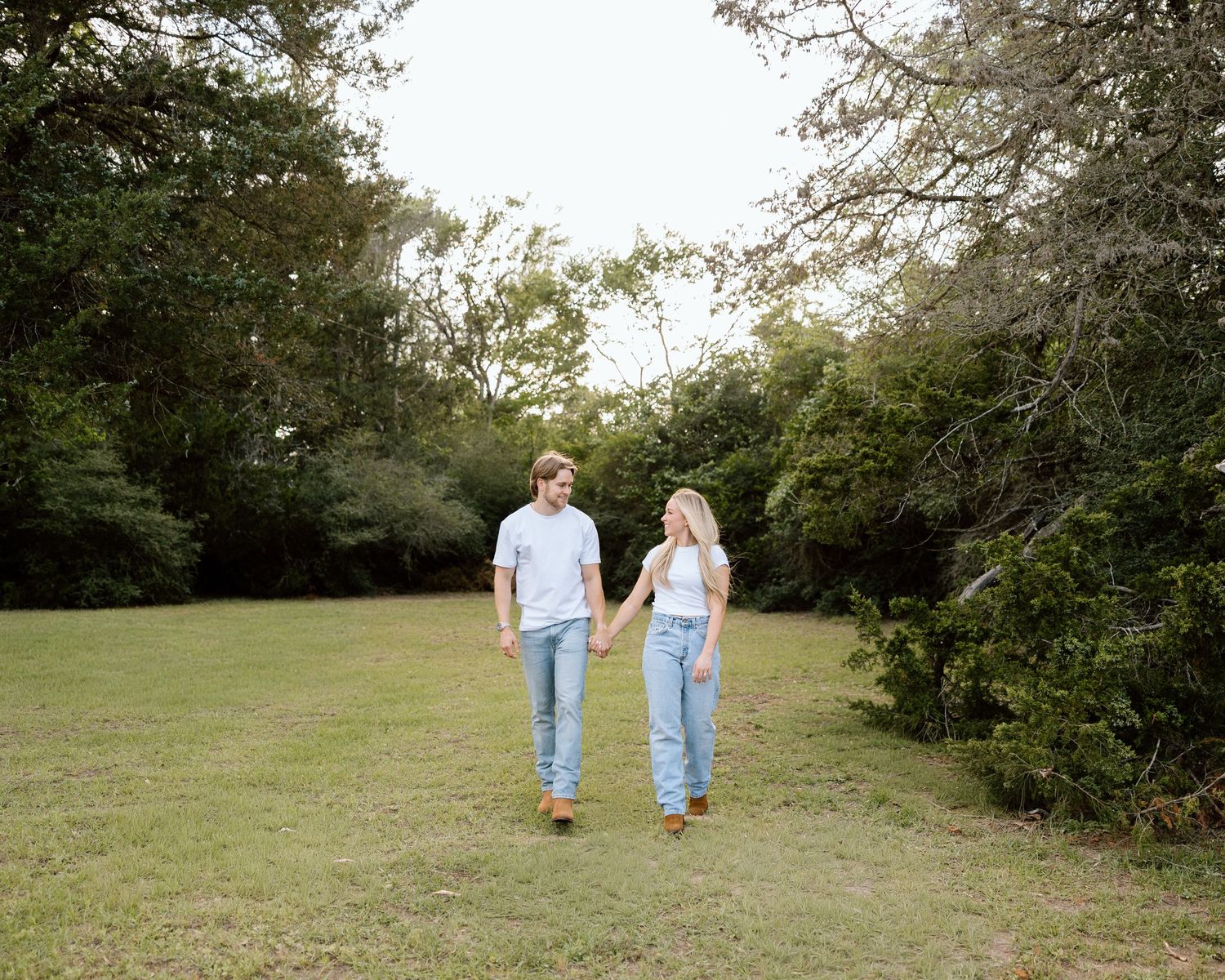 Couple holding hands walking through a lush green park surrounded by trees on a cloudy day.