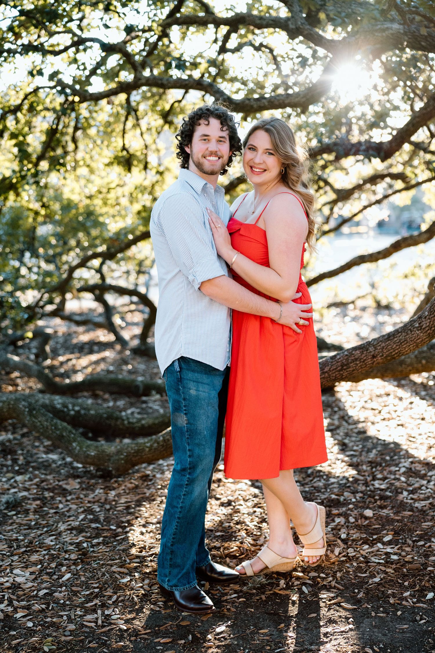 A smiling couple embraces outdoors under oak trees, woman in red dress, man in gray shirt and jeans, sunlight filtering through branches.